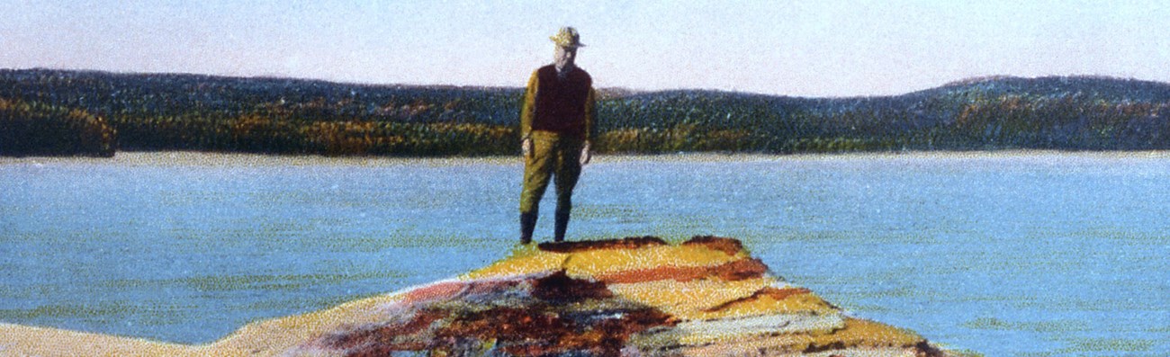 Vintage photocard showing man standing on rock next to lake