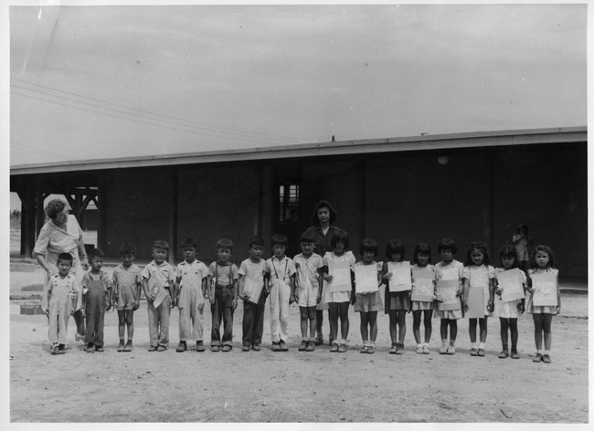 A row of Japanese American kindergarteners holds up some of their schoolwork in front of their school. Boys are at left. Girls are at right. At left a white teacher minds the kids' behavior while a Japanese American teacher stands behind the group.