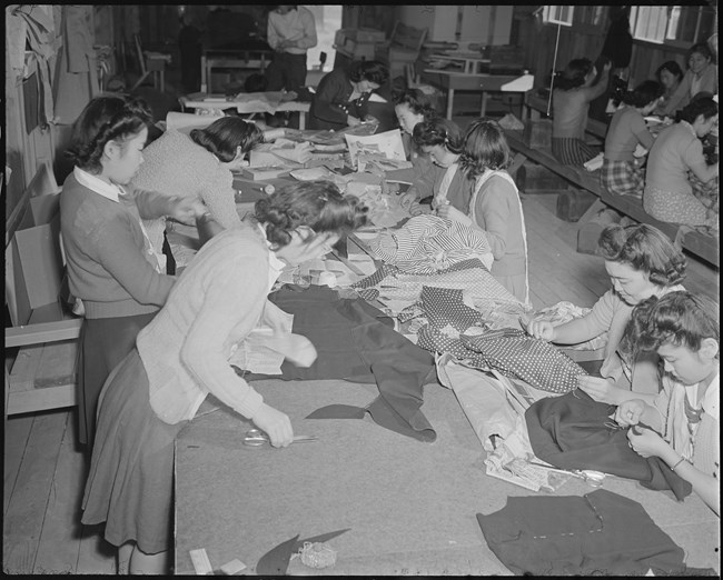 Black and white photo of young Japanese American women sewing pieces of fabric into garments at long picnic tables.