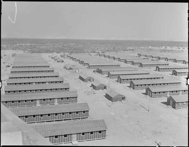 Bird’s eye view of rows of long, low barracks in a desolate landscape. The barracks are all perpendicular to a wide central road that is bisected by row of smaller buildings.