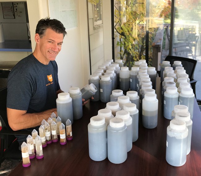Preparing insect samples Smiling man sits at a table with several plastic liter water containers filled with dark liquid.