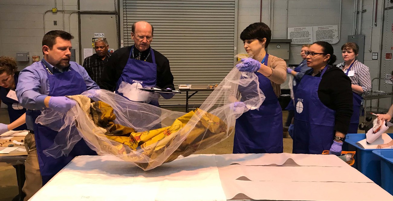 Two preservationists lifting a flood-damaged textile onto a work surface using a mesh sheet as support