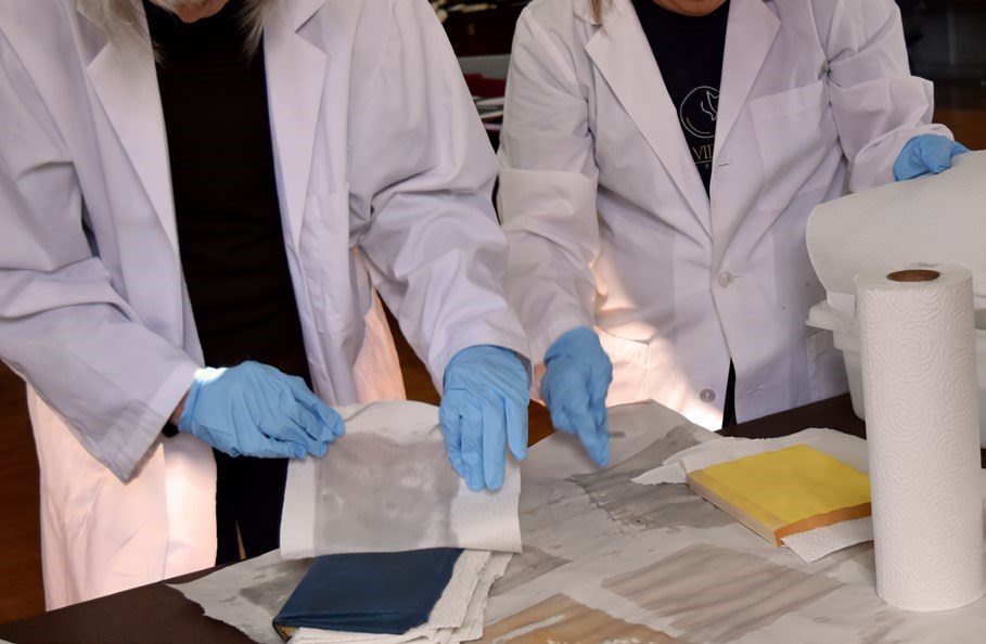 Two preservation professionals are shown placing paper towels between the pages of two wet books. Two preservation professionals are shown placing paper towels between the pages of two wet books.