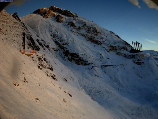 A snow-covered mountainside with the road slumping in between two construction zones, where steel bracing for a portion of the bridge is visible. A snow-covered mountainside with the road slumping in between two construction zones, where steel bracing for a portion of the bridge is visible.