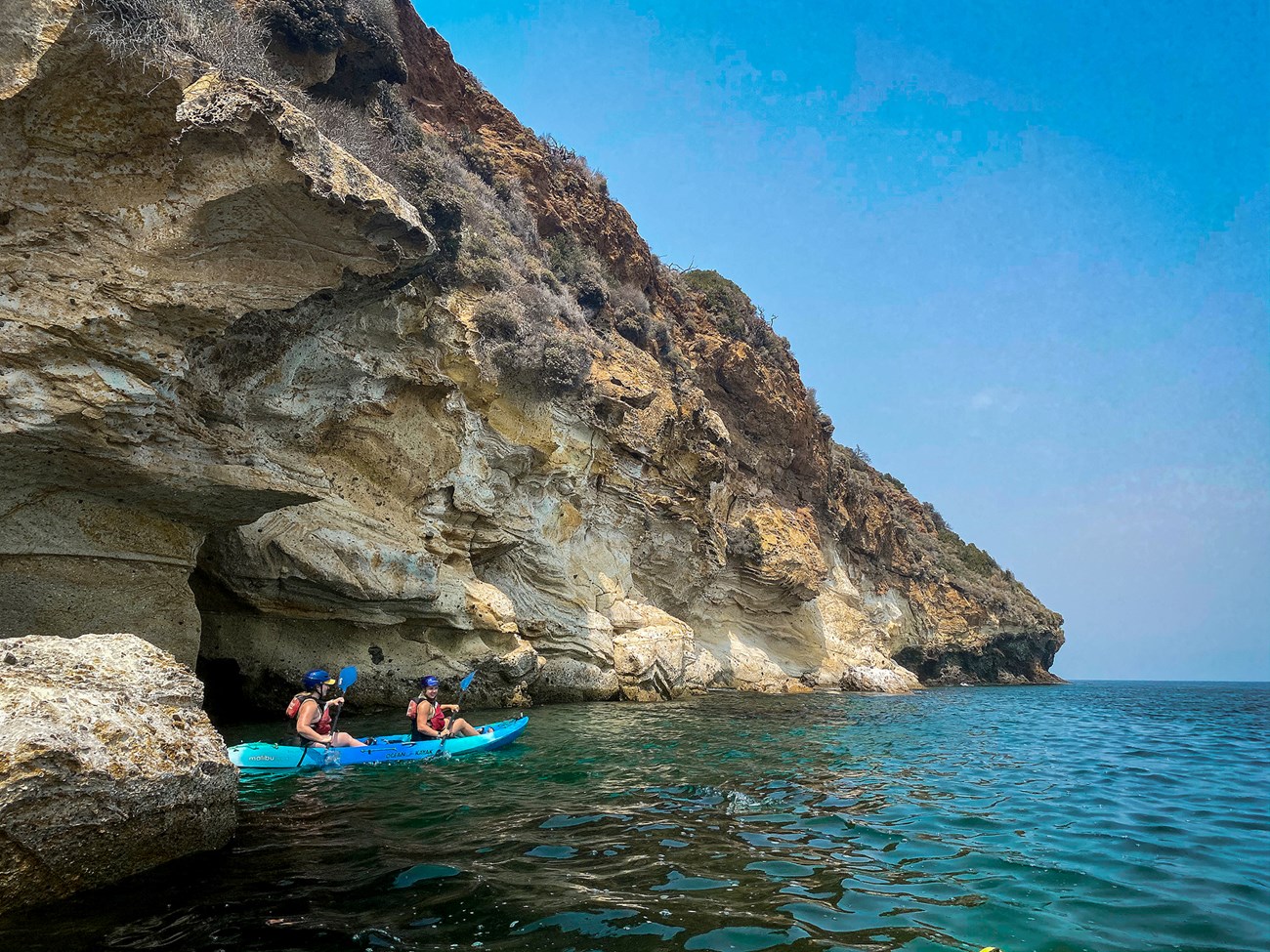 kayakers paddle along ocean cliffs with visible layering