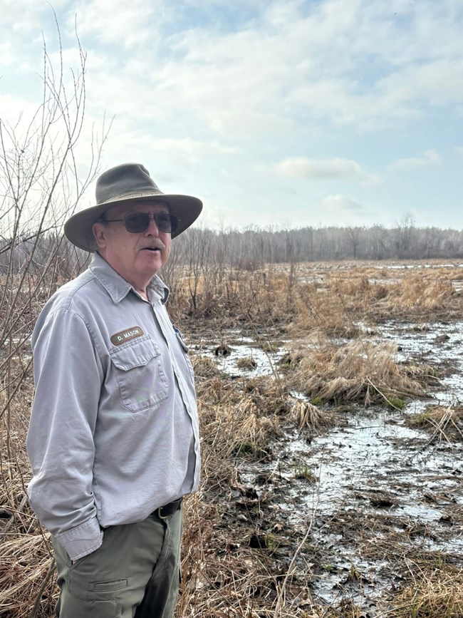 A uniformed person wearing a green wide-brimmed hat, dark-tinted sunglasses and a gray trimmed moustache, standing in front of an expansive landscape of pools of water covering dense clumps of tan-hued grasses.