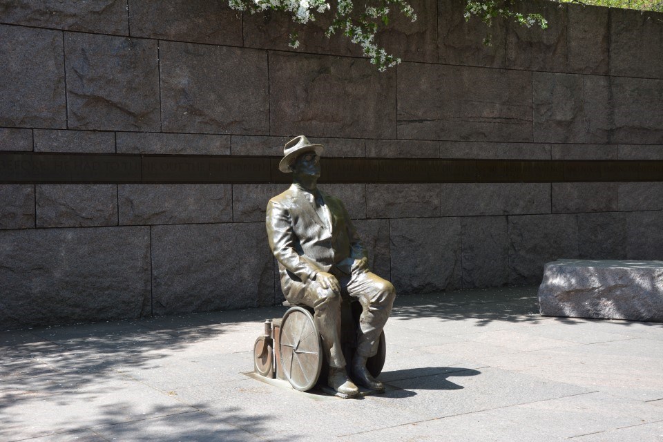 Bronze Statue of man wearing glasses, a suit, and a hat. He is seated in a wheelchair. His hands are in his lap.