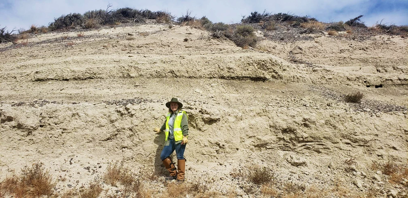 Famous Hagerman Horse Quarry site Woman wearing a wide brimmed hat, gloves, a yellow safety vest, and gaiters leaning against a bluff consisting of layers of lose, light-colored rock.
