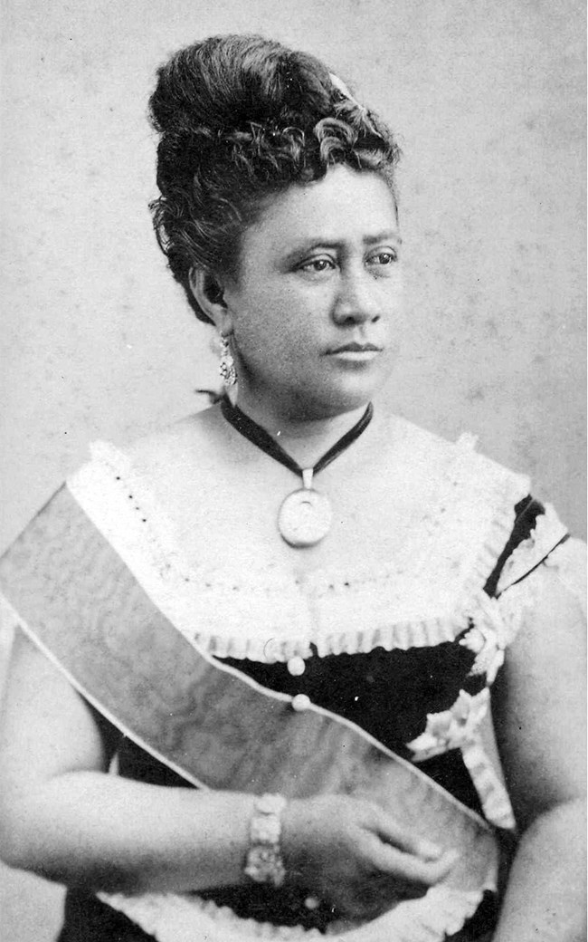 Regal Hawaiian woman in fine gown, royal sash and coiffed hair gazes into distance