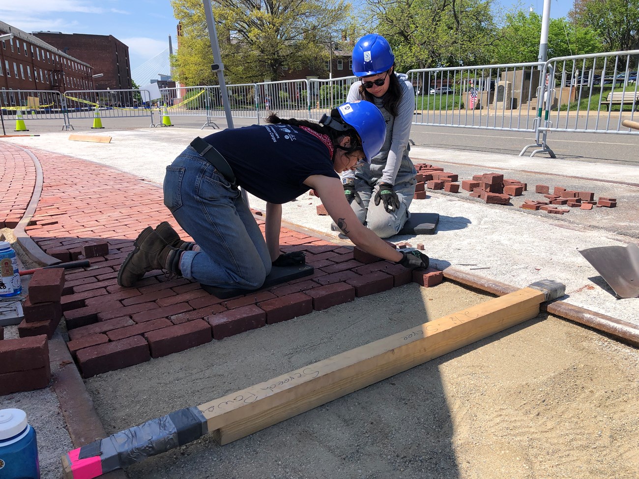 Two preservation corps members wearing hard hats and gloves. They are kneeling next to a set of trolley rails, and placing bricks between the rails to be mortared later.