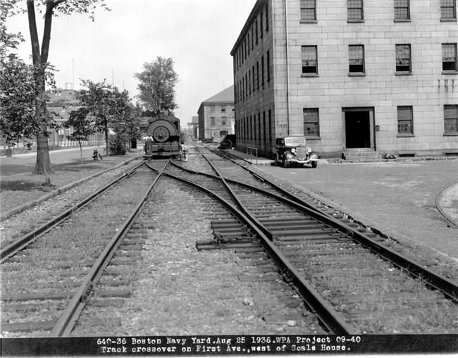 A black and white image of two parallel sets of train track receding into the background. The track on the right crosses over to the one on the left.