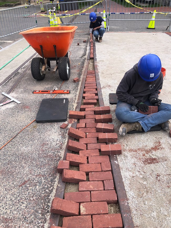 A thin strip of brick laid between two close areas of trolley track. Some bricks are partially leaning on the ground outside the track with lines, some of which have lines drawn to denote where to cut them to fit.