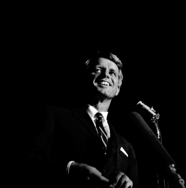 A black and white photo of RFK in a suit.  He is smiling and looking upward.