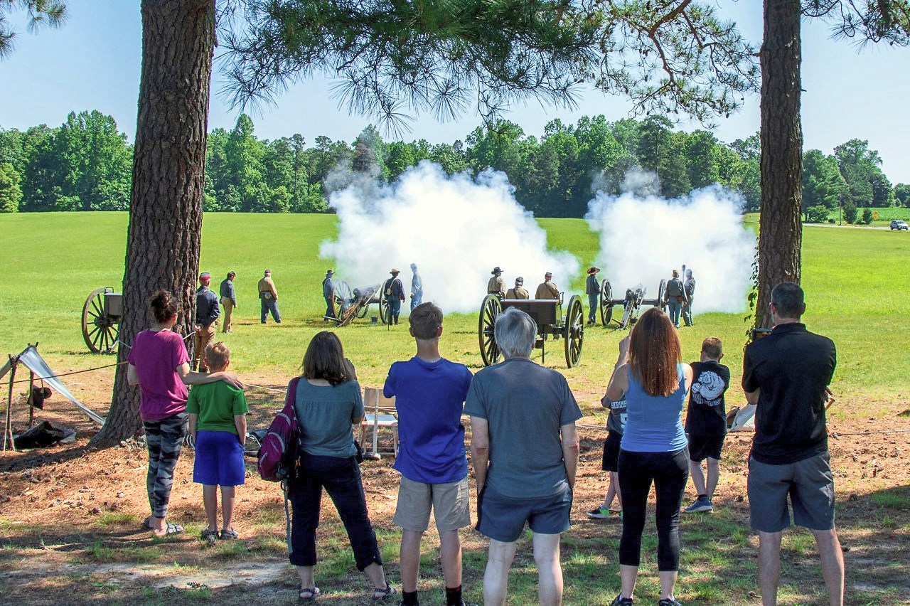 Group of adults and children under a pair of trees, some photographing/filming with mobile devices as clouds of smoke engulf two just-discharged cannons nearby.
