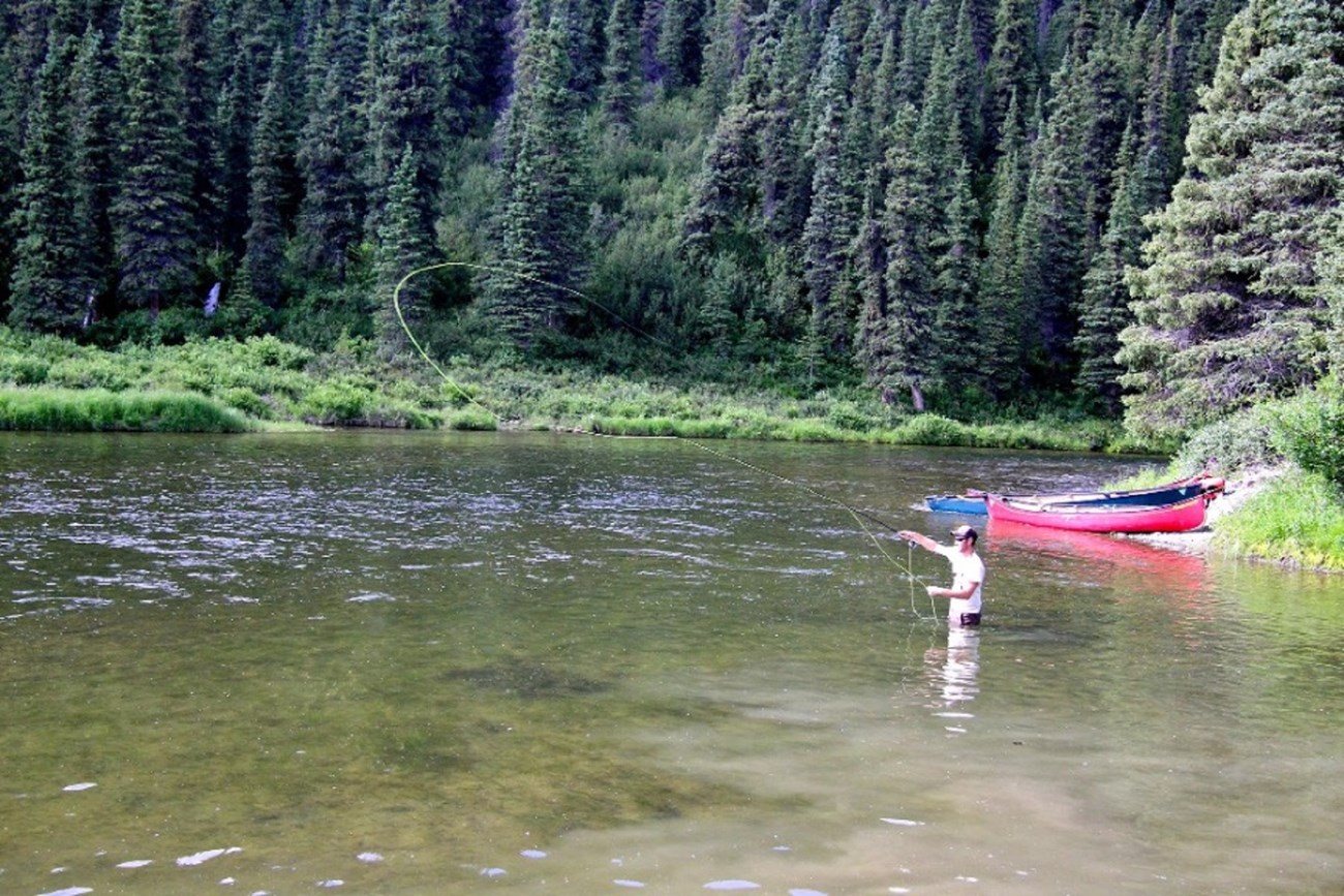 A man stands waist deep in an Alaskan river while fly fishing. Two canoes are parked on the river bank behind the fisherman