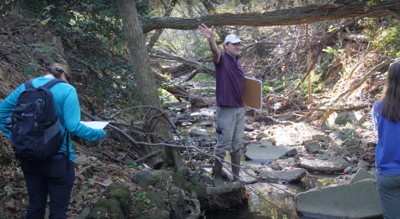 A man stands on the edge of a stream holding a white board while teaching. Two students face him.