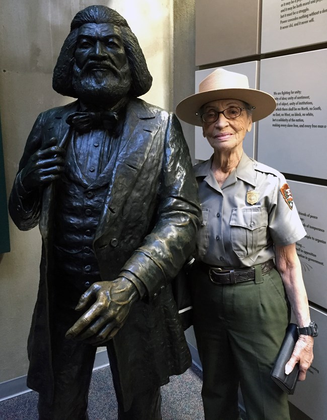 Older Black woman in NPS uniform and flathat stands proudly next to statue of Frederick Douglass