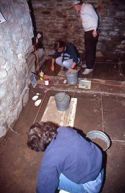 A person in a blue shirt works on the dirt floor inside a stone building. Around them are several gray buckets and archaeological tools.