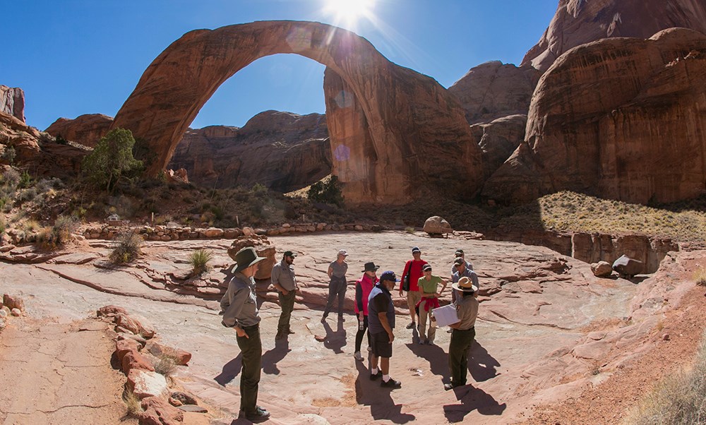 Wide view of sandstone arch with a few people standing underneath looking at park ranger