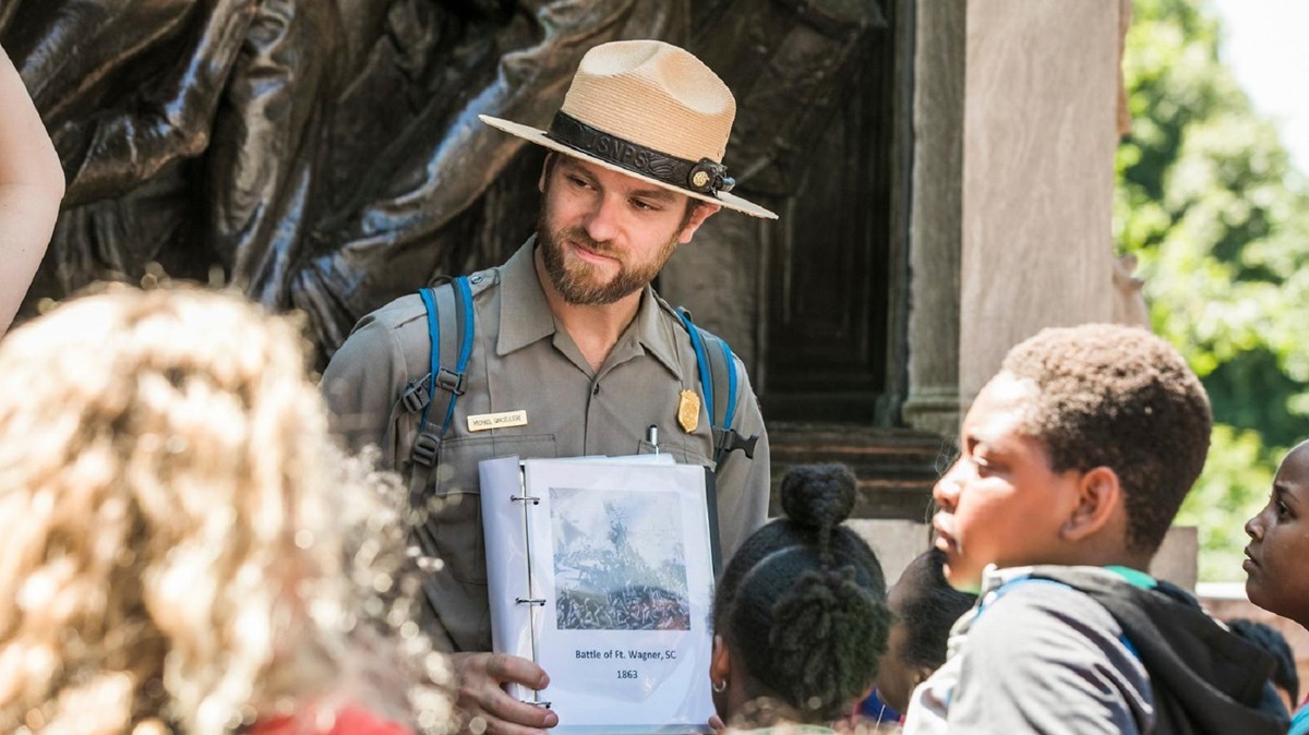 A Ranger in uniform with a flat hat stands in front of the Robert Gould Shaw Massachusetts 54th Regiment Memorial and holds up an open binder to a group of young people.