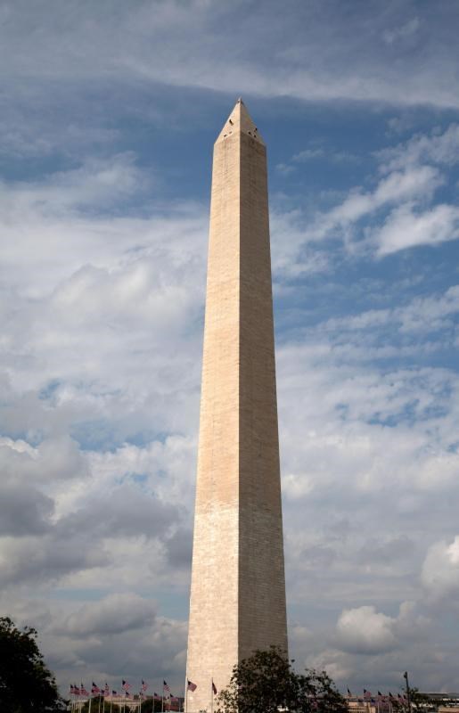 The tall stone Washington Monument obelisk is encircled by American flags and rises high above a grassy park.