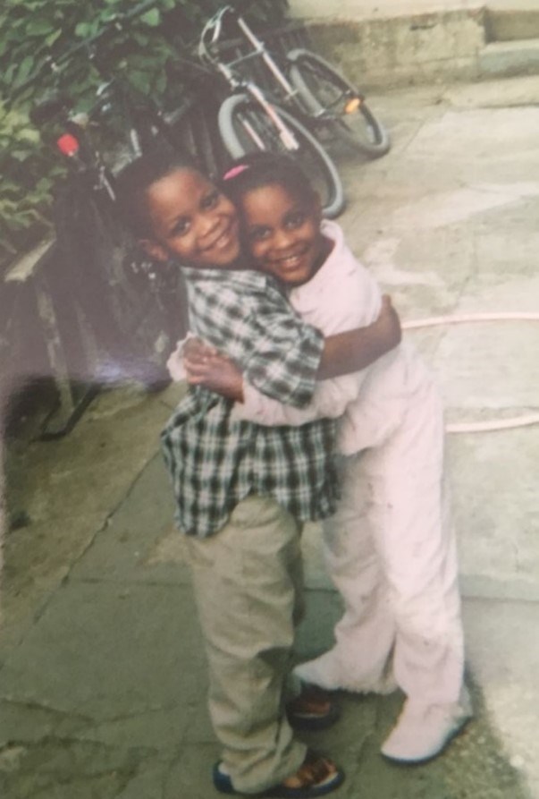 Rashaun and twin sister together in their childhood backyard Two young siblings in an embrace while standing on a paved path with a bicycle and trees in the background to the left.