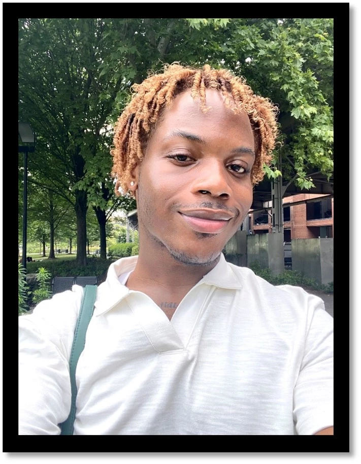 Rashaun at Georgetown Waterfront Park soaking up the sun A young individual with short brown dreadlocks, brown shirt, and green trees in the background.