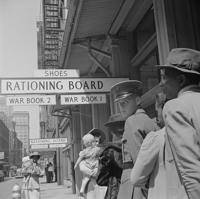 Black and white photo of white and African American people. A young man in uniform looks over his shoulder; 2 women hold young children. There are separate offices for War Books 1 and 2 and Shoes; Gasoline and Tires; and for Institutions and Restaurants.