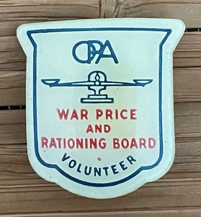 A shield-shaped white plastic pin printed in blue and red. In blue: “OPA [image of a scale] VOLUNTEER.” In red: “WAR PRICE AND RATIONING BOARD.” Collection of the author.