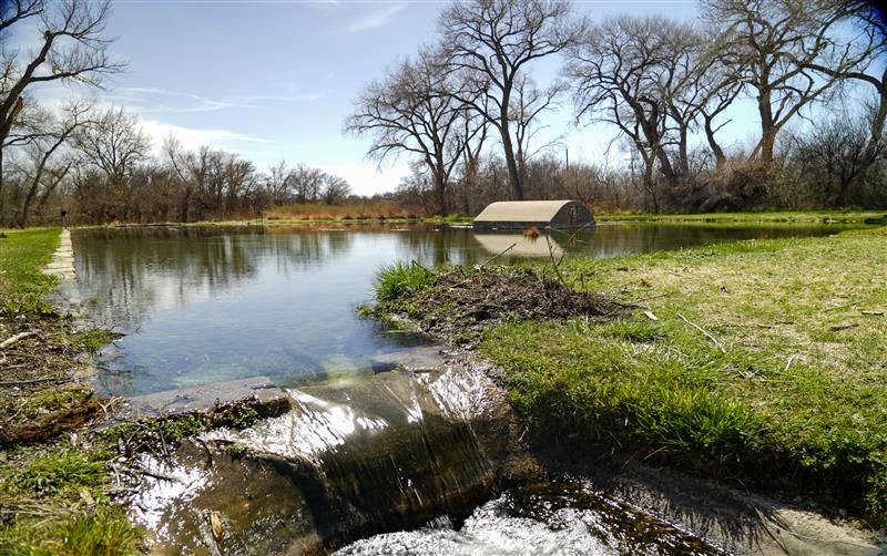 View of a small pond with submerged gray building, trees, and blue skies in the background. The pond flows over a small built waterfall in the foreground.