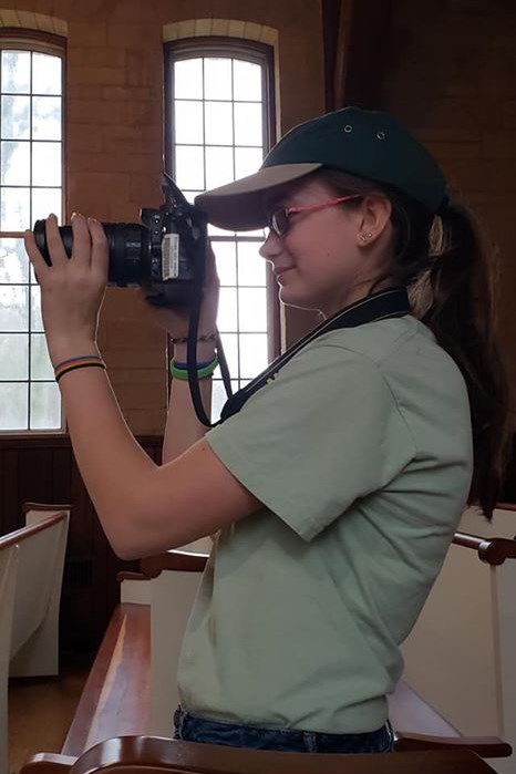 Rebecca Snider taking pictures for Lincoln Day Side profile of a girl in a baseball cap looking through the viewfinder of a DSLR camera.