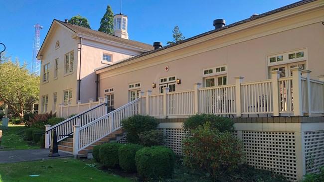 Red Cross Vancouver Barracks A photo of a light-colored building on a sunny day. The building has a porch in front of it with stairs leading up to a main entrance. Over the entrance is a small plaque with a red cross.