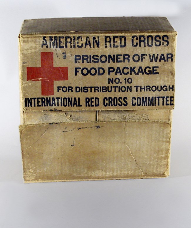 Brown cardboard box, marked with a red cross and lettered in black “American Red Cross Prisoner of War Food Package No. 10 for Distribution through the International Red Cross Committee.”