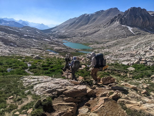 Lake monitoring crew hiking toward Guitar Lake. Three people carrying backpacks hike down a rocky slope toward a mountain lake.