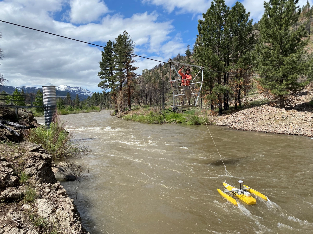 Collecting streamflow measurements Person in a metal cage suspended from a cable over a river, holding on to a floating yellow flow measurement device via a long cable.