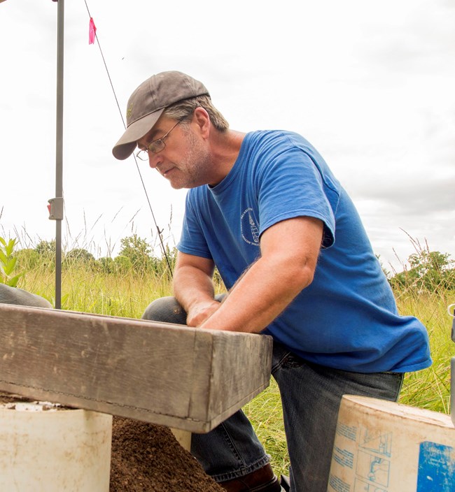 Man in ballcap and glasses bends over a wood tray above a pile of soil. Tall grass and treeline in background.