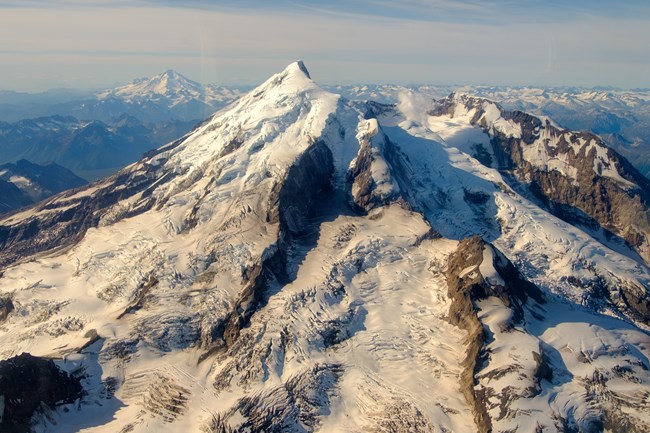 View of Redoubt in 2018, Iliamna is visible on the horizon to the left. View of two snow capped volcanoes and clear skies.