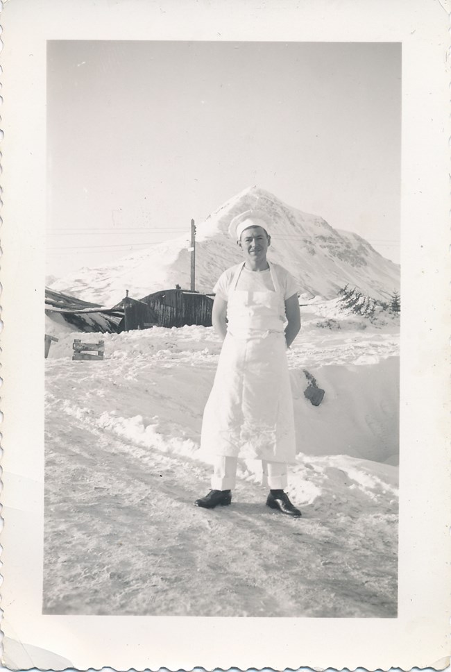 Man in white chef's uniform standing on a snowy road, in front of a white tent and a large, snow covered mountain in the background.