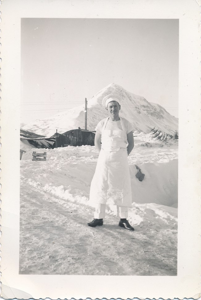 RCAF chef in Kodiak, Alaska Man in white chef's uniform standing on a snowy road, in front of a white tent and a large, snow covered mountain in the background.