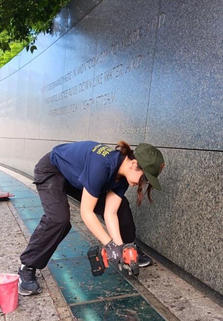 Woman in blue shirt, black pants, and a green hat uses a power tool. She is surrounded by large stone slabs and tiles that are part of a monument