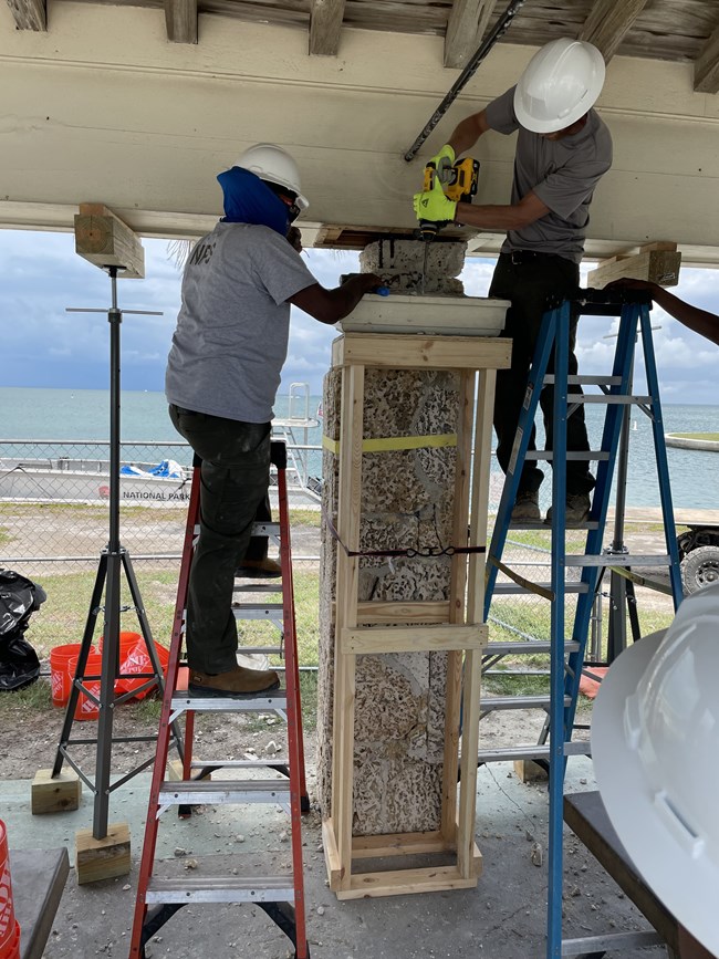Removing and Dismantling Column Crown Boca Chita Pavilion Two NPS employees in hardhats are outdoors, standing on ladders, assisting one another (one with a drill, the other with a hand tool) in column repair work.