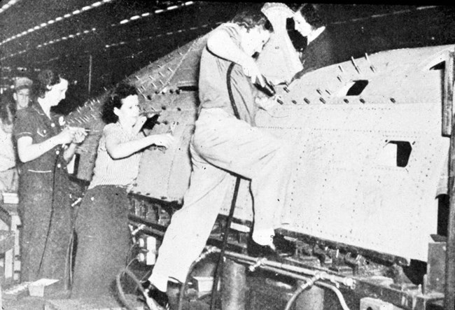 A white woman steps up onto a ledge to drive rivets into the side of a plane. In the background, other women predrill holes, reach into the plane’s body, or work from inside the cockpit.