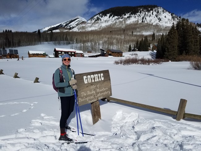 Person on cross-country skis stands in a snowy landscape next to a sign for the town of Gothic, Colorado.