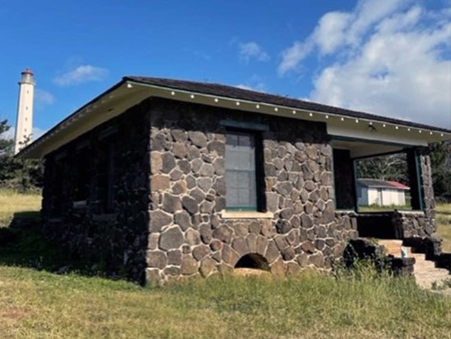 Neatly restored house made of lava rock sits in grass, Molokai Light standing behind it.