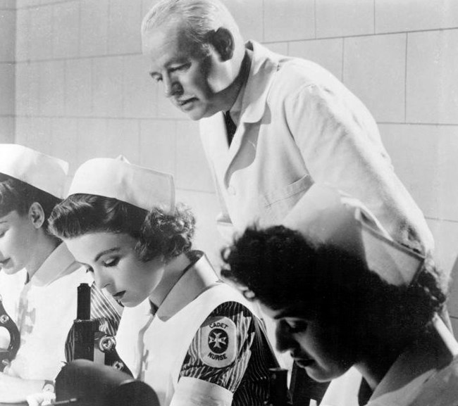 Black and white of three women in nurse caps with a Cadet Nurse Corp Patch looking into microscopes. An older man in a white coat looks over their shoulder.