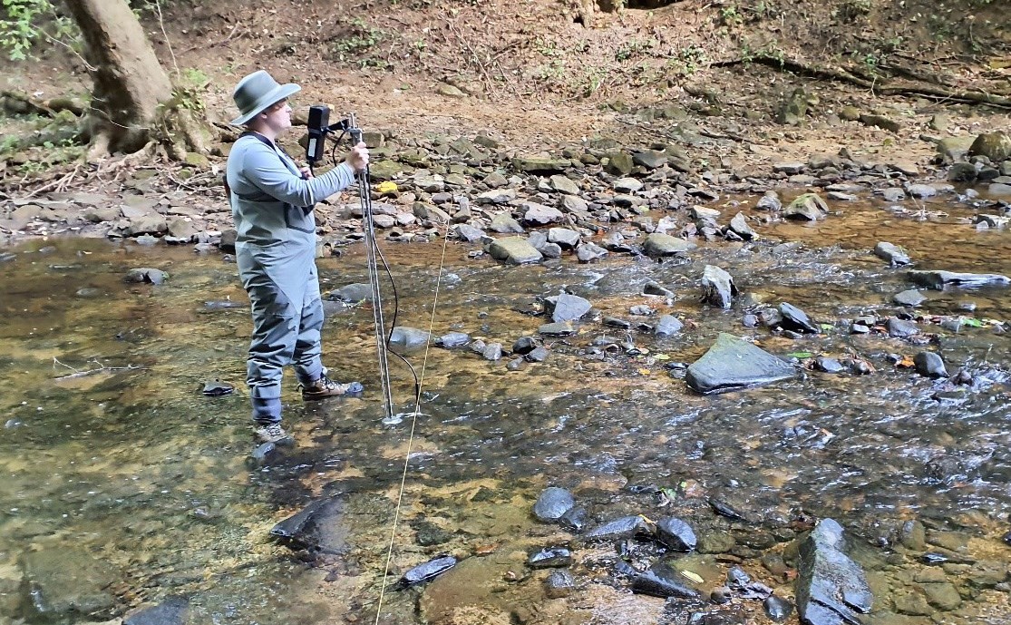 Rhea operating FlowTracker Person with a tripod standing in a shallow, rock-filled stream
