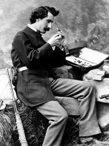 black and white photo of a seated nineteenth century man inspecting a small object in his hands. He has