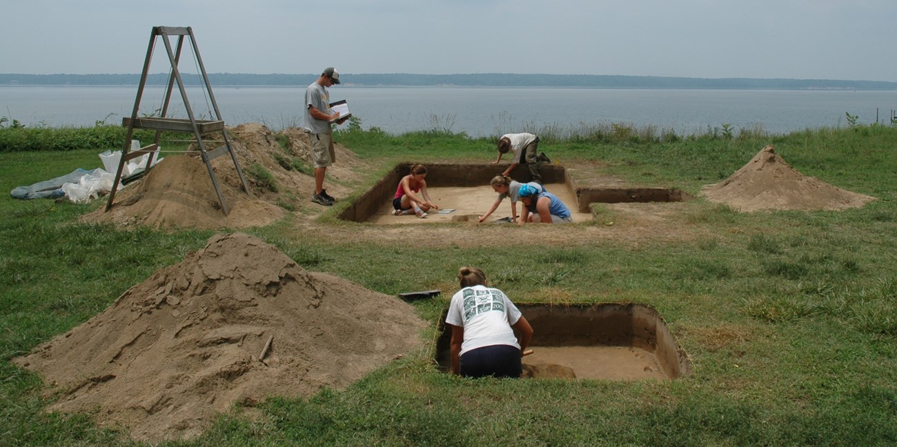 Archeologists excavate rectangular dig units on a flat lawn overlooking a wide river.