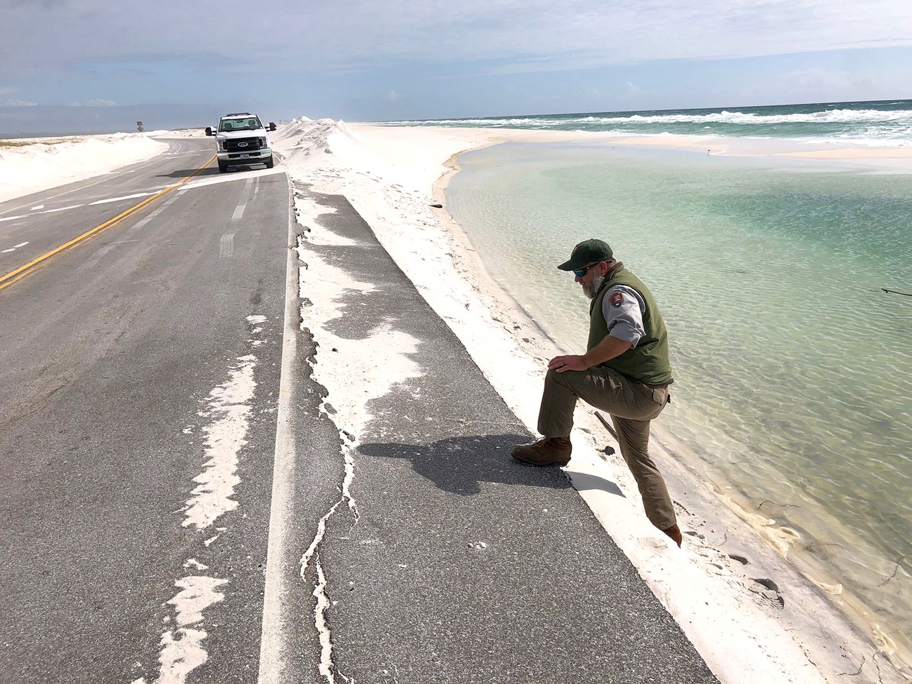 Man in NPS uniform stands with his foot on the edge of a sand-covered road next to the ocean. A truck is in the background.