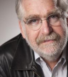 An anglo man about sixty with gray hair, beard, and glasses, wearing a leather jacket over a pinstriped dress shirt.